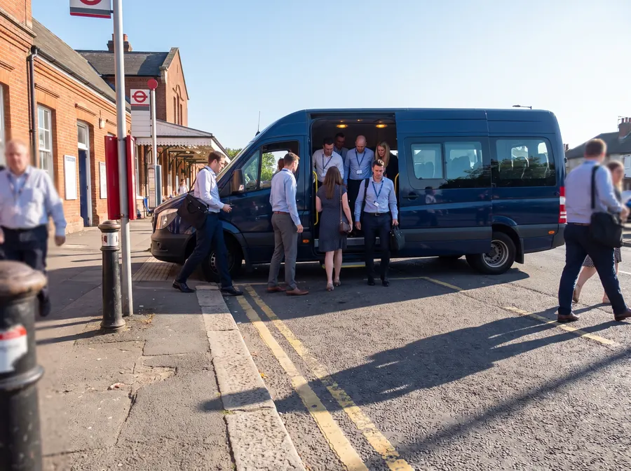 A dark navy 16-seat minibus parked at the kerb on the road outside Welling railway station on a bright clear summer morning, strong sunlight and crisp shadows, a corporate group in business-casual clothes with laptop bags and lanyards boarding through the open door